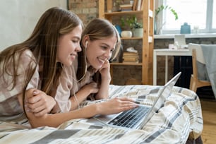 Süße lächelnde Teenager-Zwillinge mit Kopfhörern und Laptop, die Online-Filme ansehen, während sie sich am Wochenende im Bett ausruhen