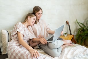 Two smiling restful girls with laptop watching movie or communicating with parents or friends in video chat while lying on bed