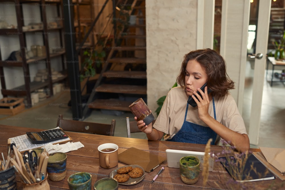 Young female potter sitting at desk with tools and handmade mugs and discussing order with buyer by phone