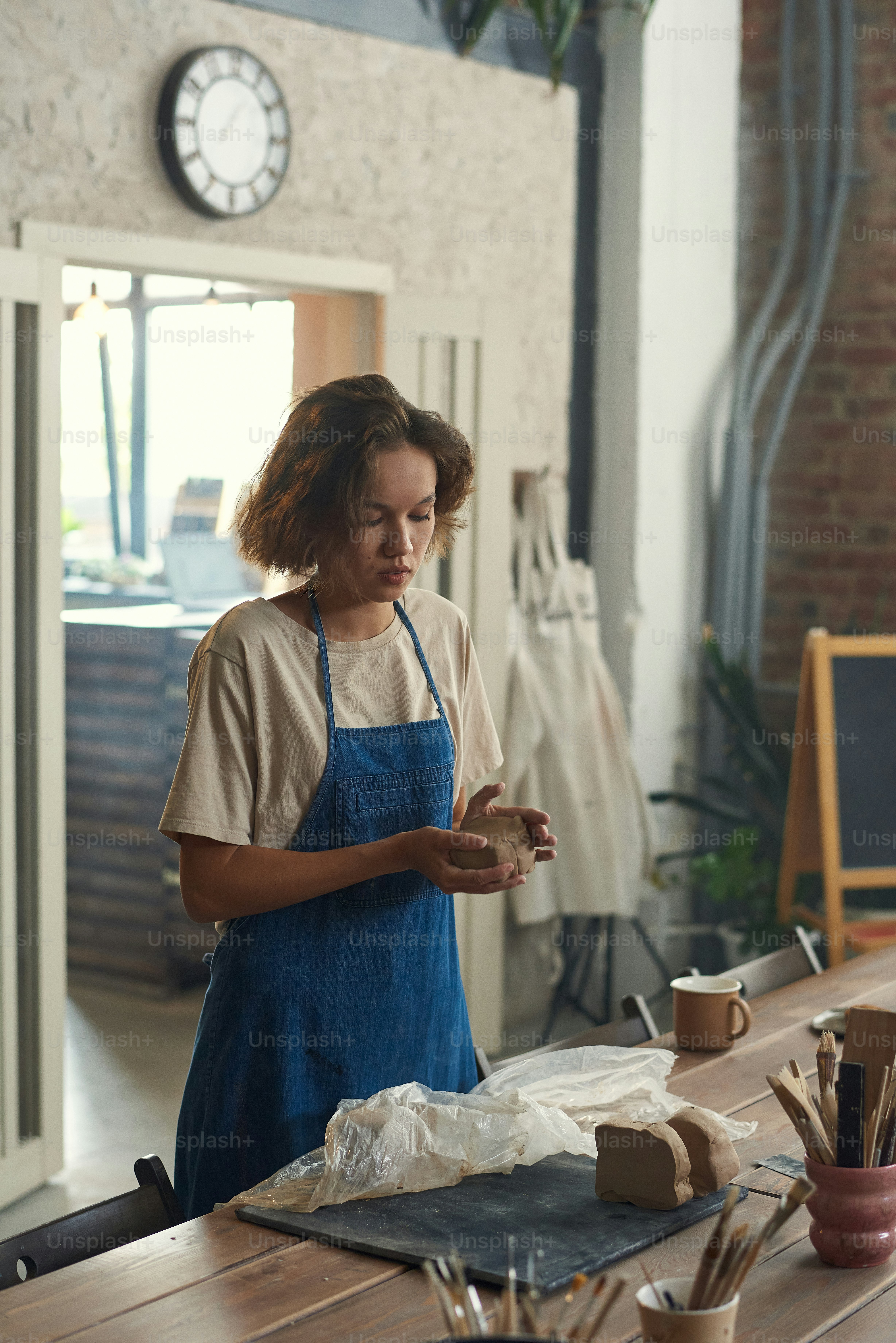 Serious young Caucasian woman in apron standing at wooden table with craft tools and molding clay with hands