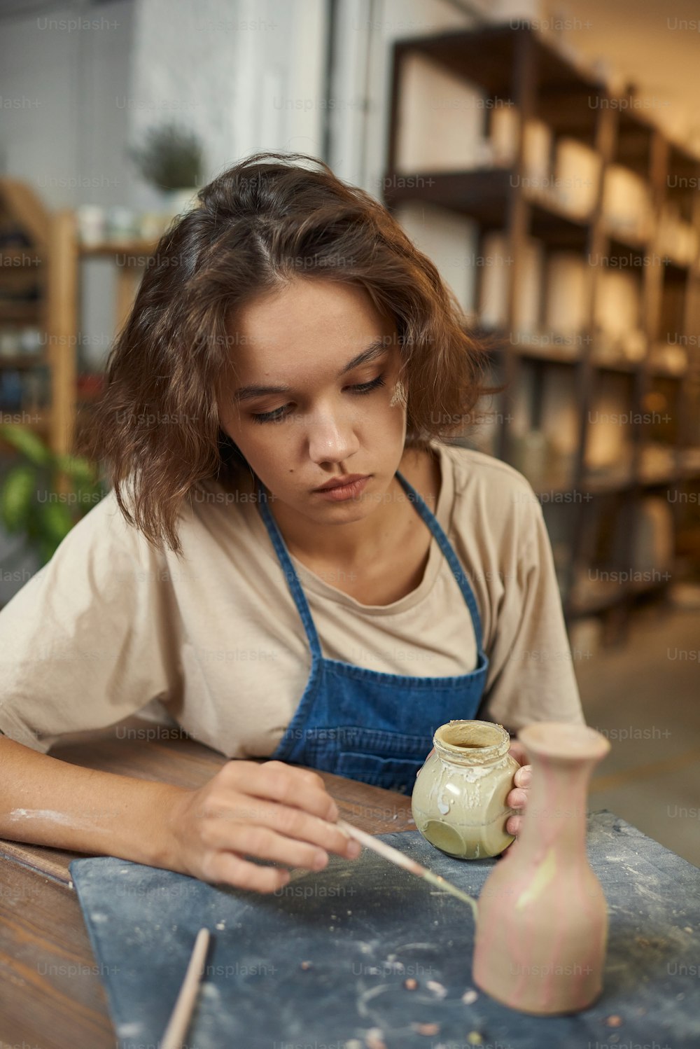 Concentrated young Caucasian potter sitting at table and painting vase while working on its design in workshop