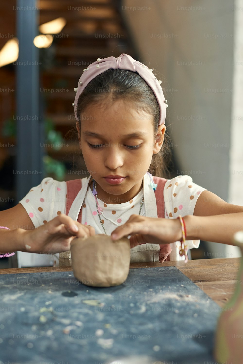 Concentrated curious mixed race girl in headband sitting at table and sculpting clay at school class in pottery workshop