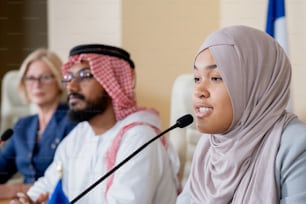 Attraktive junge muslimische Frau im Hijab spricht in das Mikrofon, während sie an einer Konferenzdiskussion teilnimmt