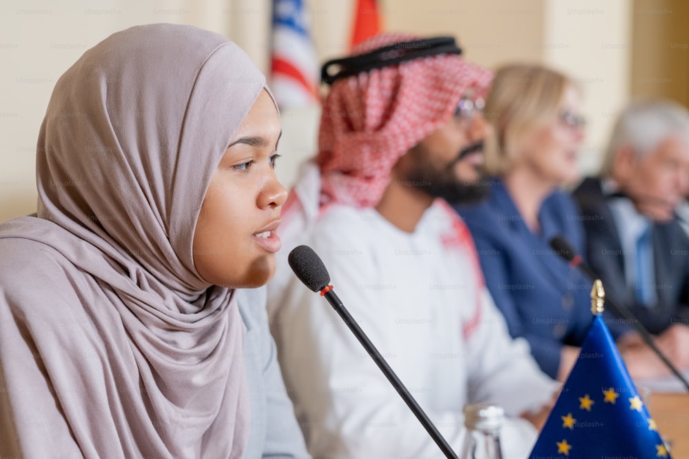 Confident young Muslim female politician in hijab speaking into microphone while nswering question of journalist