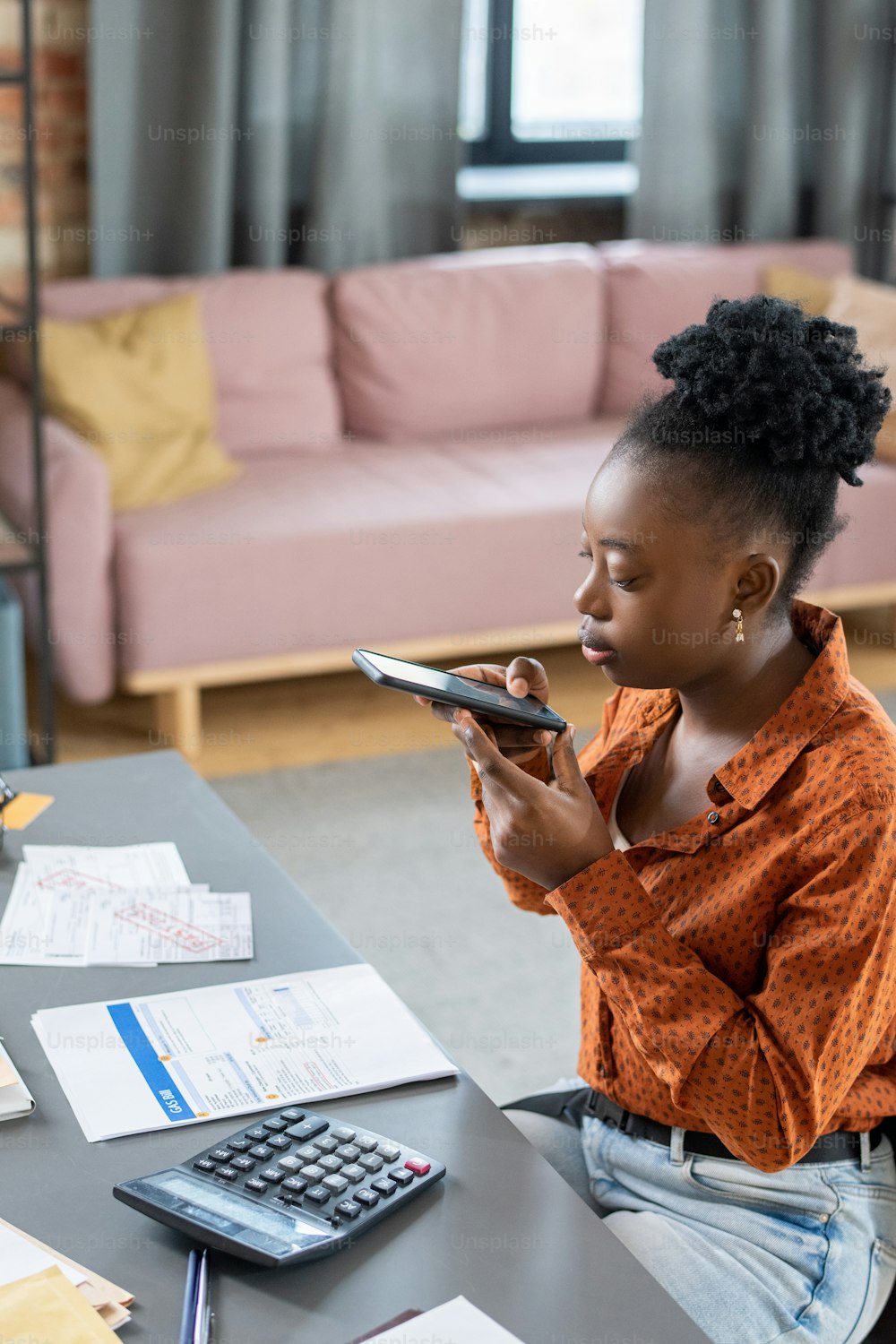 Serious young African American woman sitting at table with calculator and photographing tax document on phone