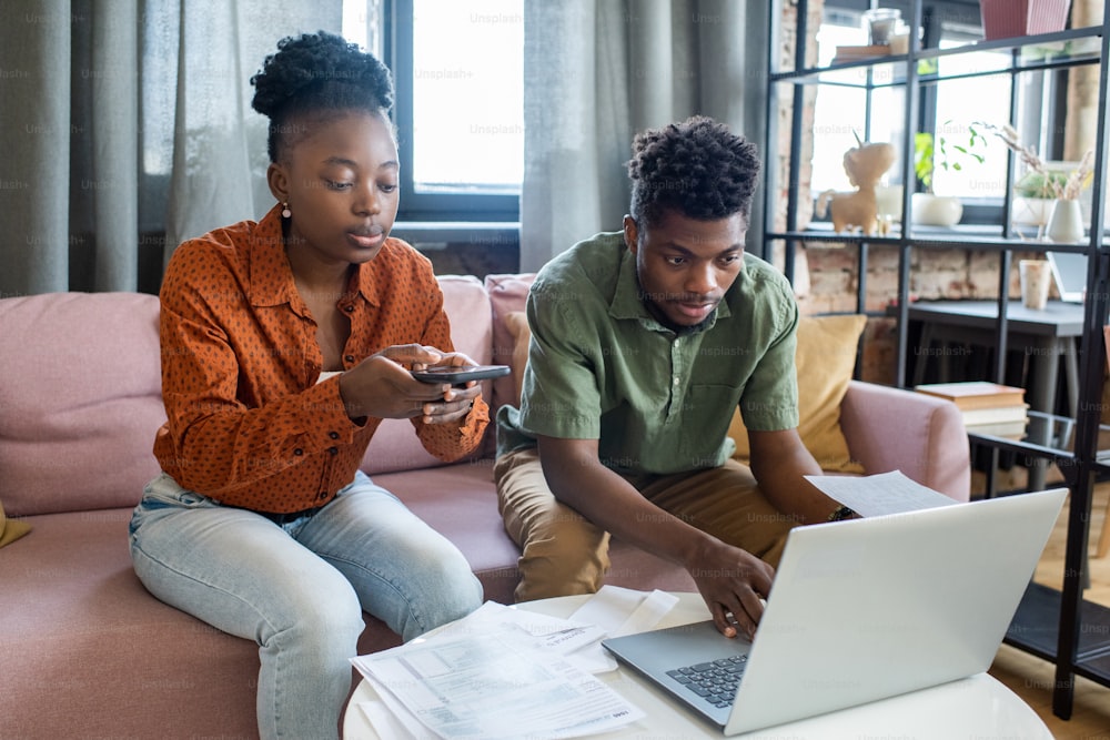Joven pareja afroamericana con trajes casuales usando dispositivos modernos mientras prepara documentos de impuestos