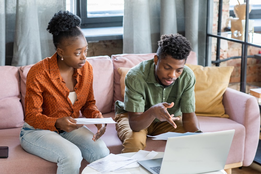 Joven afroamericano serio sentado en una mesa de café con computadora portátil y examinando las finanzas con su novia