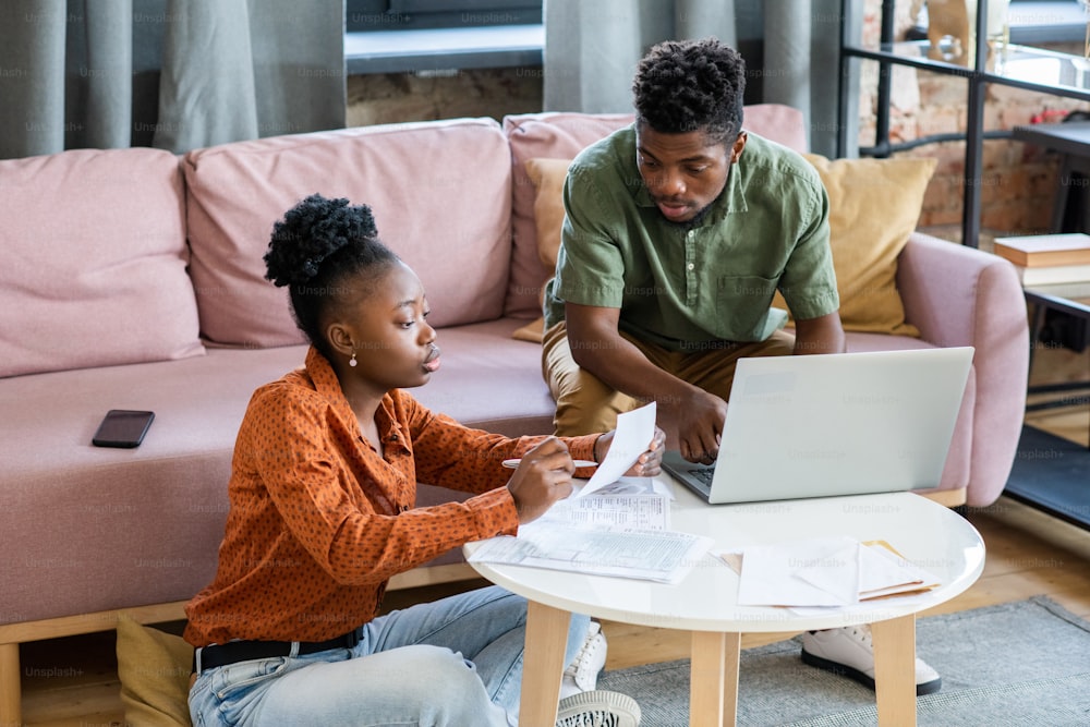 Ernstes junges afroamerikanisches Paar, das mit Papieren und Laptop am Kaffeetisch sitzt und über Finanzen diskutiert