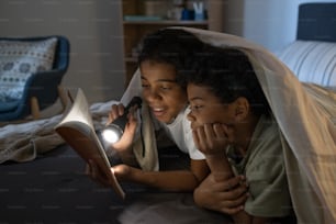 Positive African American girl shining flashlight on book while reading it to brother in darkness
