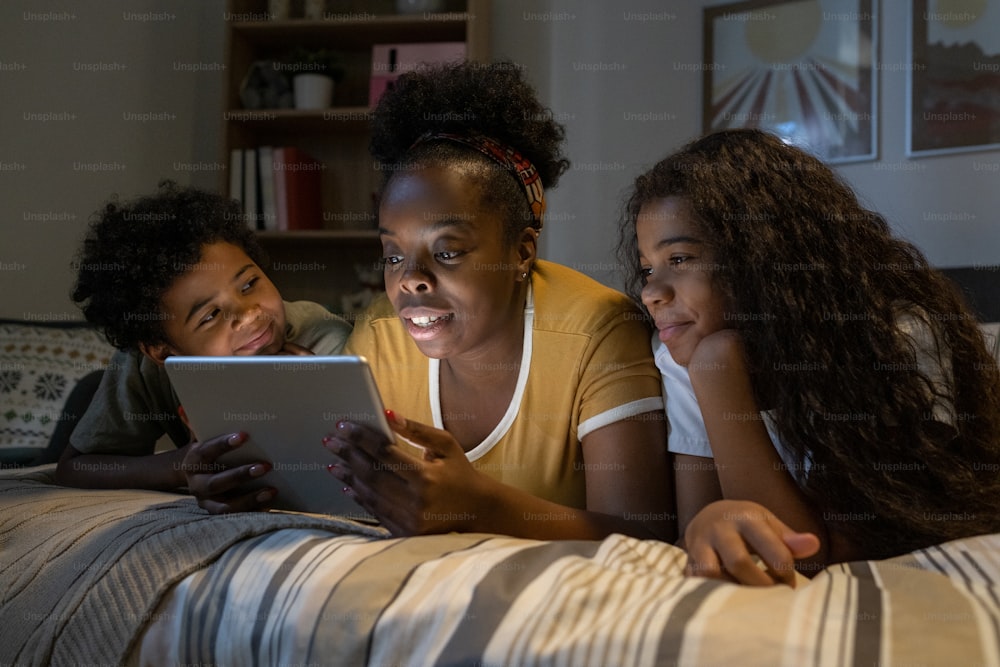 Careful middle-aged African American mother lying on bed and using tablet while reading online book to children in dark room
