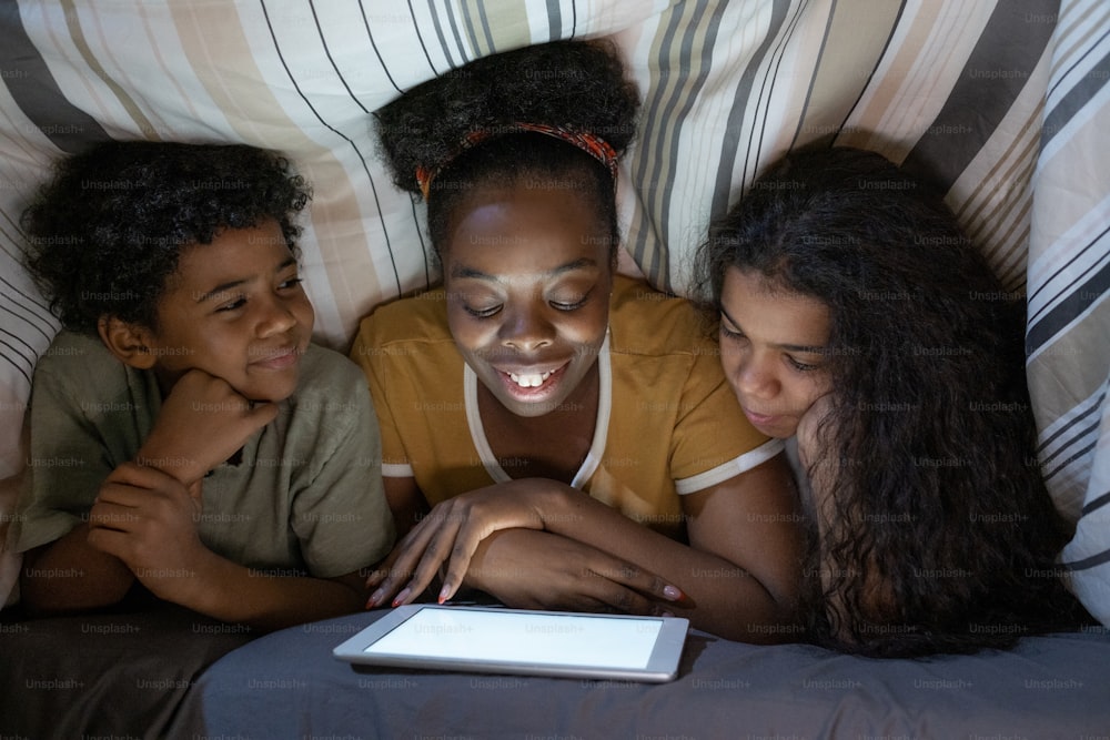 Smiling young African American mother lying on front and reading book to children under duvet