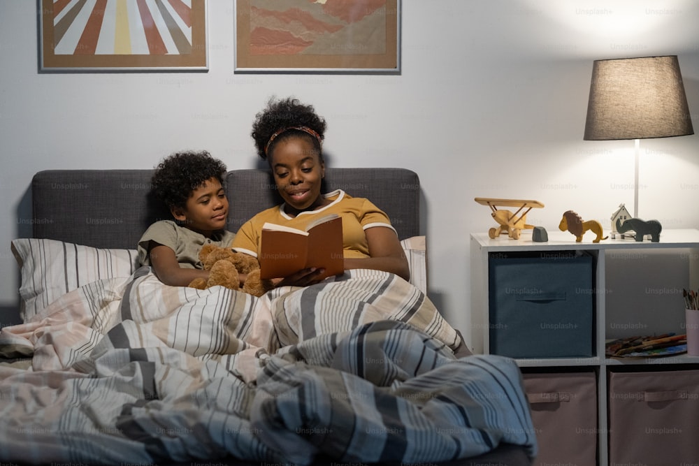 Smiling African American mother with curly hair sitting in bed and reading book to cute son in nursery room