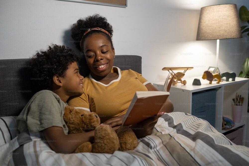Happy young African American mother in homewear sitting in bed and reading book while smiling at son