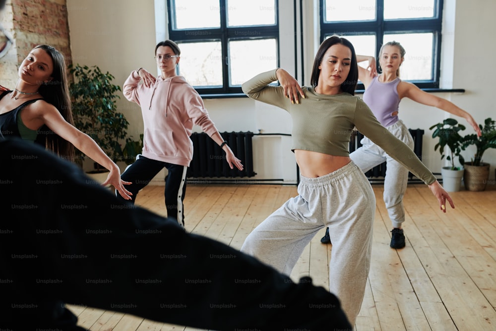 Chicas adolescentes y chico en ropa deportiva repitiendo ejercicio de baile después de su instructor o líder durante el entrenamiento en el amplio estudio loft