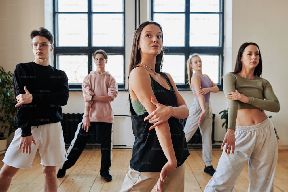 Group of contemporary teens in pants and tanktops repeating after dance instructor while learning new movements of vogue dancing