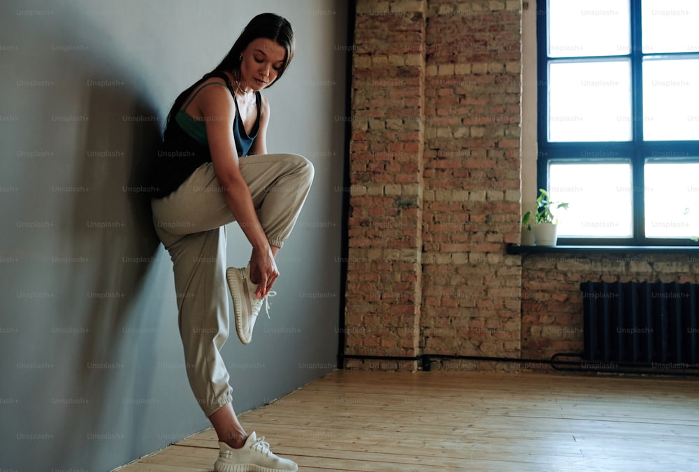 Contemporary young female dancer in activewear tying shoelace before repetition while standing against grey wall in loft studio