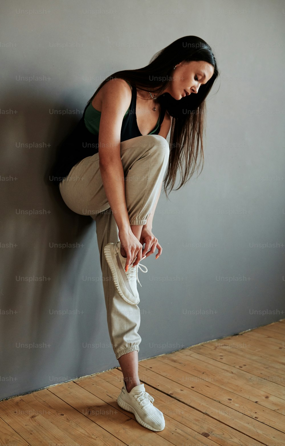 Giovane performer femminile in pantaloni e canottiera che lega il laccio delle scarpe su scarpe da ginnastica bianche mentre si trova contro il muro grigio nello studio del loft