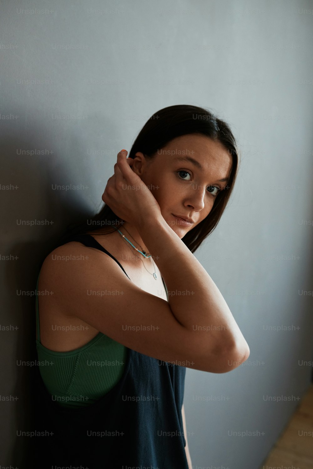 Young contemporary woman in black tanktop touching her dark long hair and looking at camera while standing by grey wall of studio