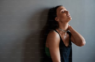 Junge müde Vogue-Tänzerin oder andere Darstellerin in schwarzem Tanktop, die in der Pause an der grauen Wand des Loft-Studios steht und sich ausruht