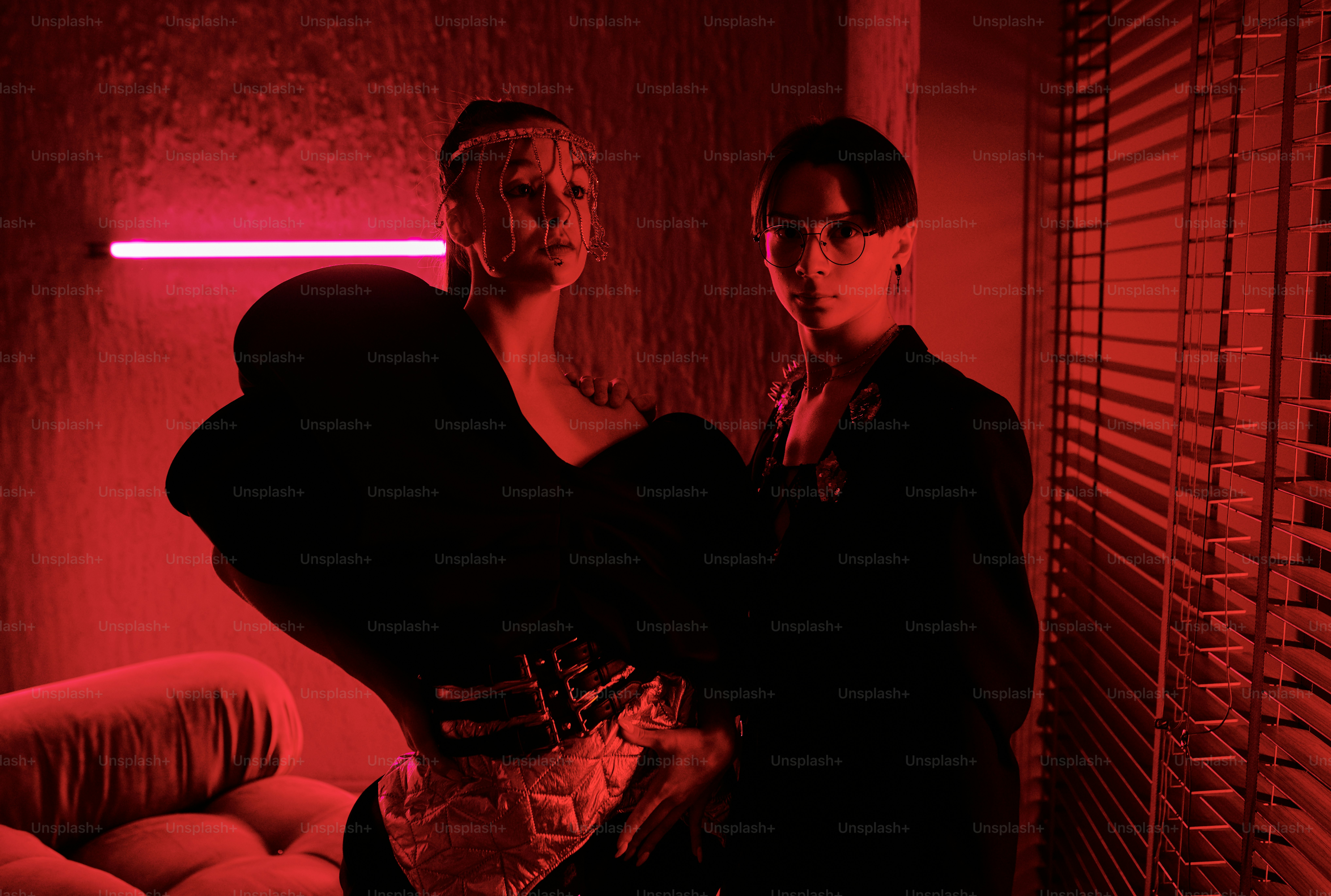 Two young vogue dancers in posh stage costumes giving performance in studio of modern night club lit with dark red light of neon lamps