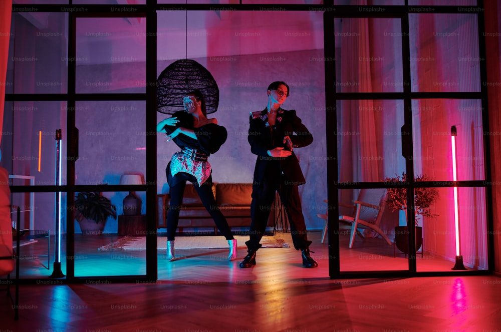 Dos adolescentes dinámicos con atuendos elegantes que bailan en el estudio loft, apartamento o club nocturno iluminado por luces de neón rosas y azules
