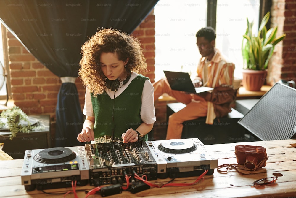 Mujer joven en ropa casual de pie junto a una mesa de madera y usando el controlador de DJ mientras crea nueva música en el ocio contra el hombre negro