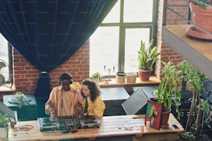 Girl with wavy hair standing close to young black man mixing sounds on dj booth while both creating new music by table in loft apartment