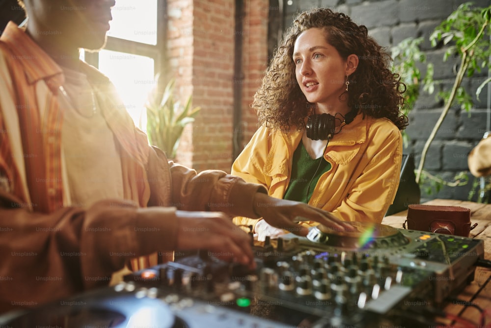 Pretty girl with dark long wavy hair talking to her African American boyfriend mixing sounds on dj set while creating new music