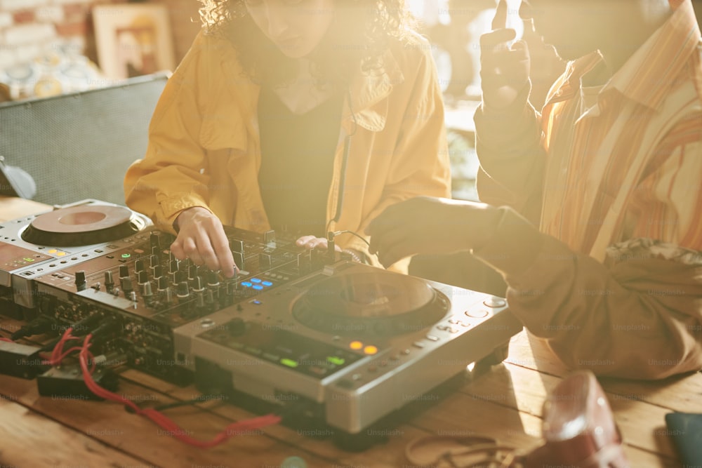 Zwei junge interkulturelle Menschen stehen mit DJ-Set am Tisch und mischen Sounds, während sie gemeinsam ihre Freizeit in einer Loft-Wohnung verbringen