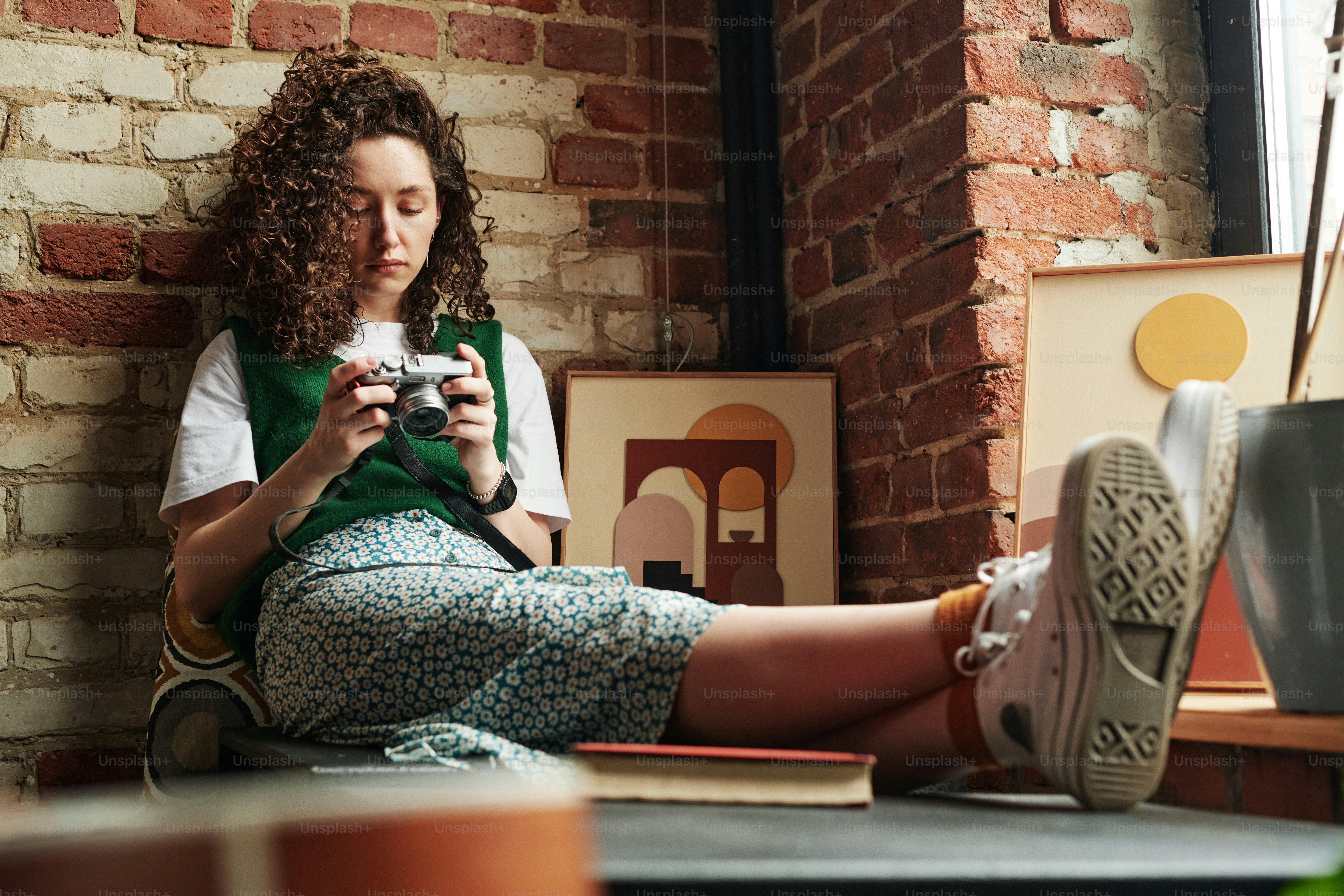 Menina repousante com cabelo ondulado escuro olhando para photocamera em suas mãos enquanto sentado pela parede de tijolos e duas fotos no apartamento loft