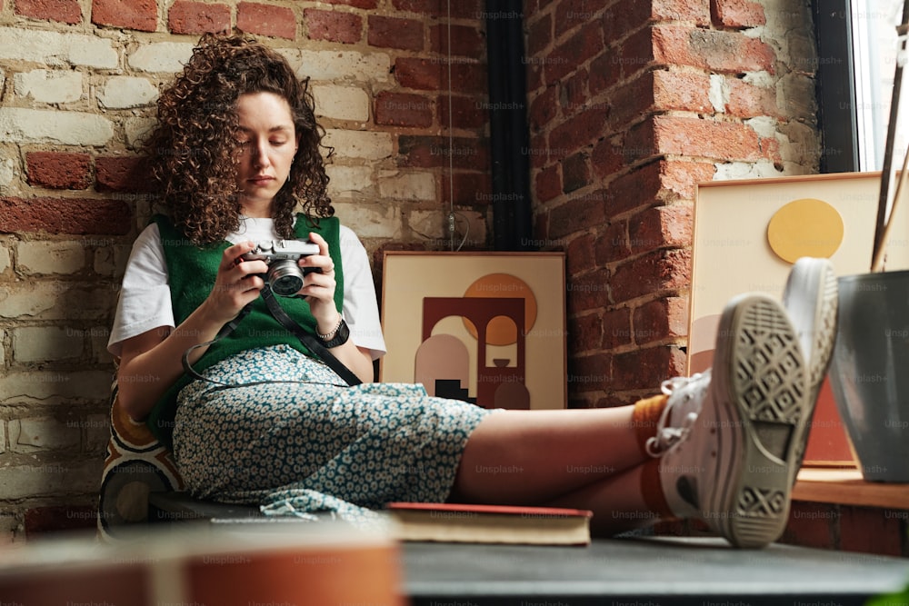 Fille reposante avec des cheveux ondulés foncés regardant un appareil photo dans ses mains tout en étant assise près d’un mur de briques et deux photos dans un loft