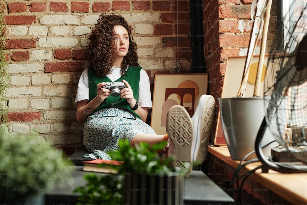 Ruhiges Mädchen mit Fotokamera in den Händen, das an der Ziegelmauer sitzt und durch das Fenster schaut, während es sich in der Loft-Wohnung ausruht