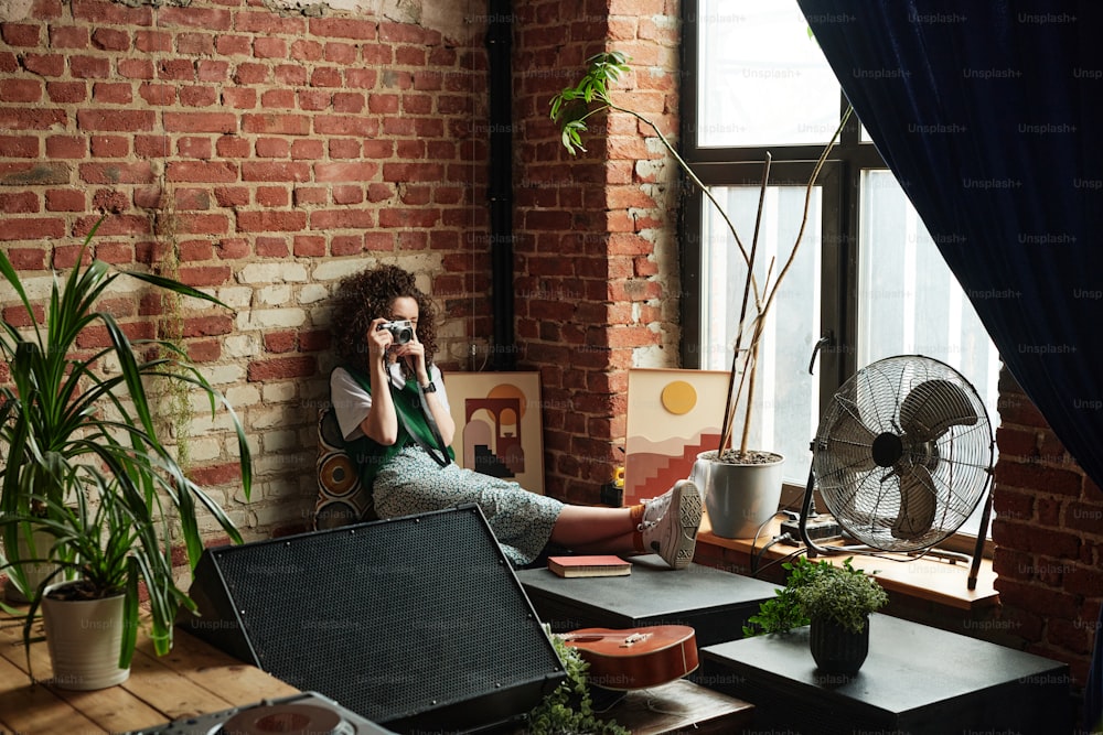 Nettes Mädchen in Freizeitkleidung, das auf der Fensterbank mit Luftstromventilator sitzt, Hauspflanze im Blumentopf und Bild im Rahmen und macht ein Foto vom Wohnzimmer
