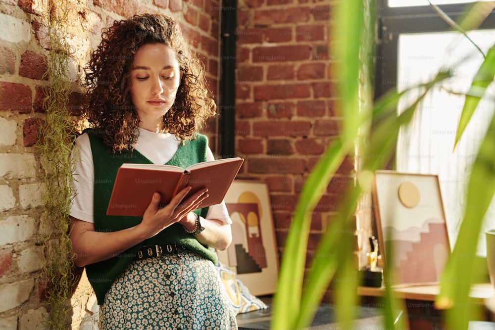 Young serious female student in casualwear holding open book and reading it at leisure while standing by brick wall in loft apartment