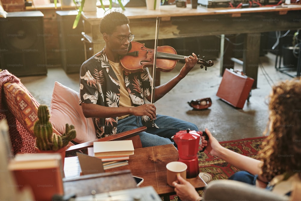 Young black man with string musical instrument performing piece of classic music in front of his girlfriend holding mug and teapot