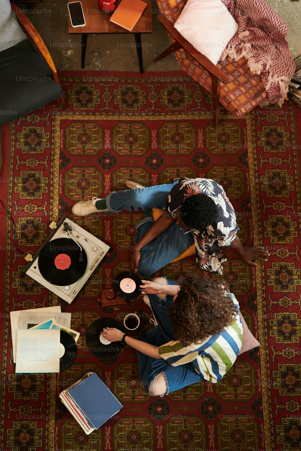 Überblick über ein junges interkulturelles Paar in stilvoller Freizeitkleidung, das auf dem roten Teppich im Wohnzimmer sitzt und Vynil-Platten auf dem Plattenspieler hört