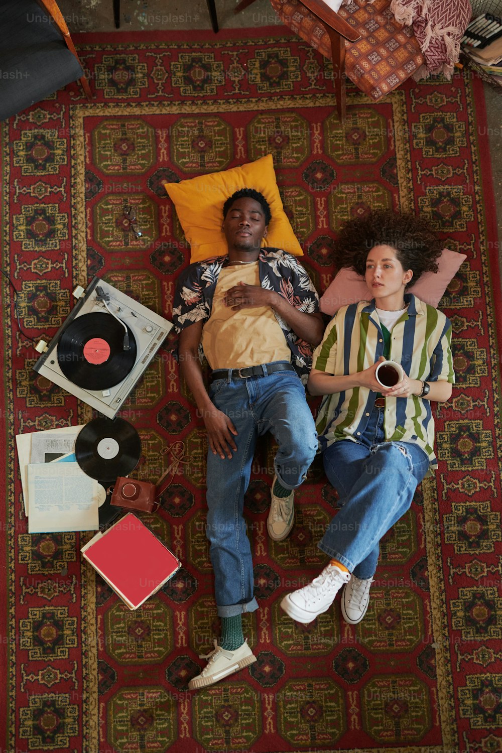 Young interracial dates keeping heads on pillows while relaxing on red carpet on the floor of living room and listening to vynil disks