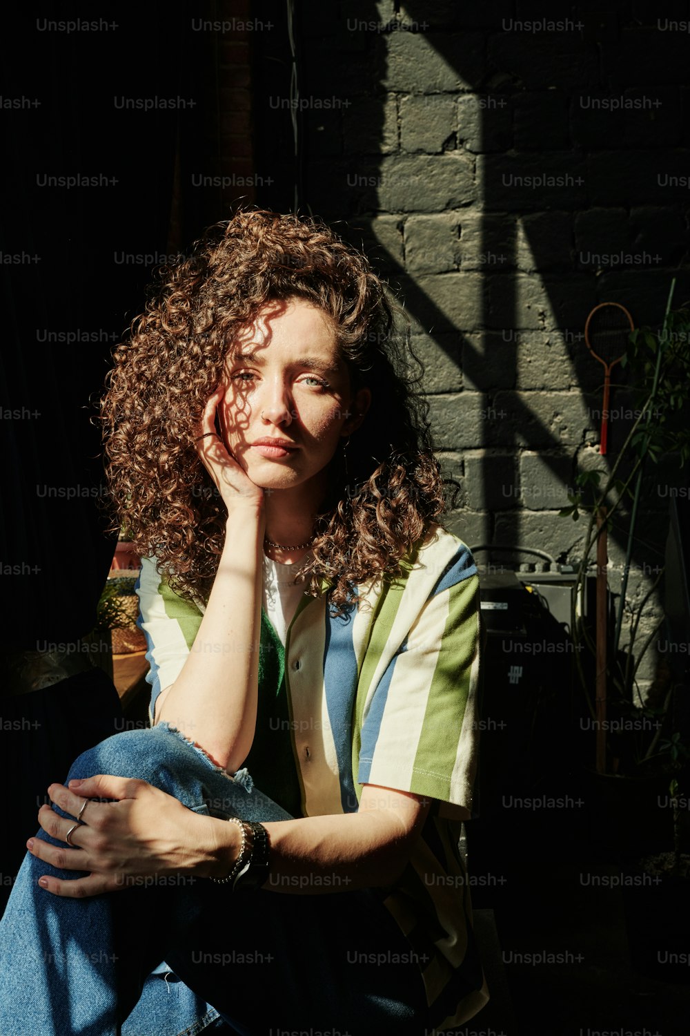 Young serene female with curly brown hair keeping hand on cheek while sitting in dark room and looking at camera against grey brisk wall