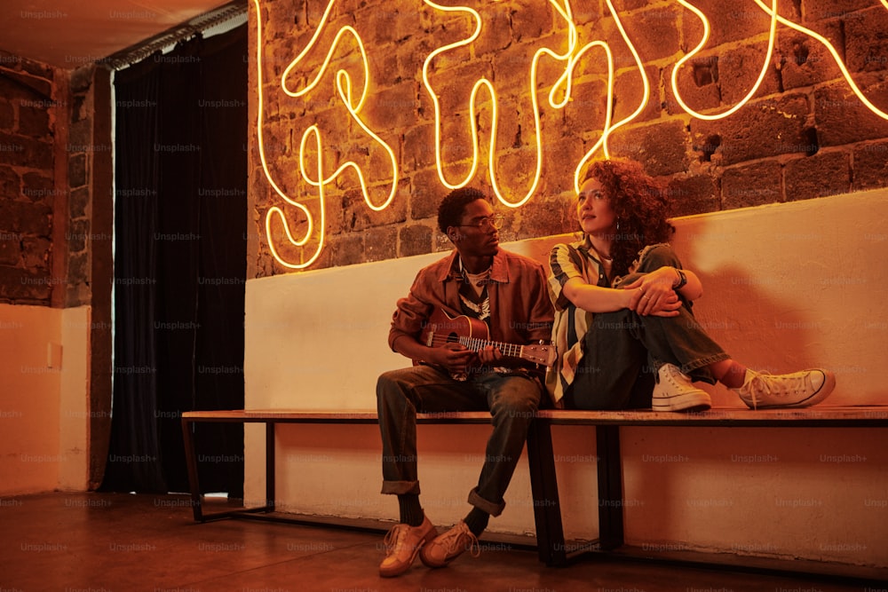 Young black man playing acoustic guitar while sitting on bench by brick wall next to his girlfriend in stylish casual apparel in studio