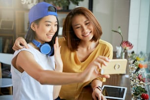 Pretty smiling Vietnamese teenage girls hugging and taking selfie in cafe