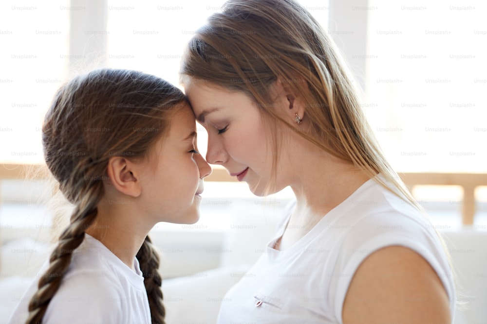Side view portrait of loving mother touching noses with cute little girl lit by serene morning sunlight, copy space