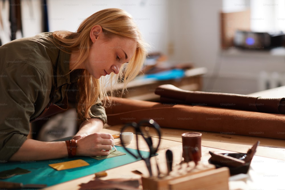신발 제작 아틀리에에서 일하는 동안 가죽 패턴을 추적하는 젊은 여성의 따뜻한 톤의 초상화, 복사 공간