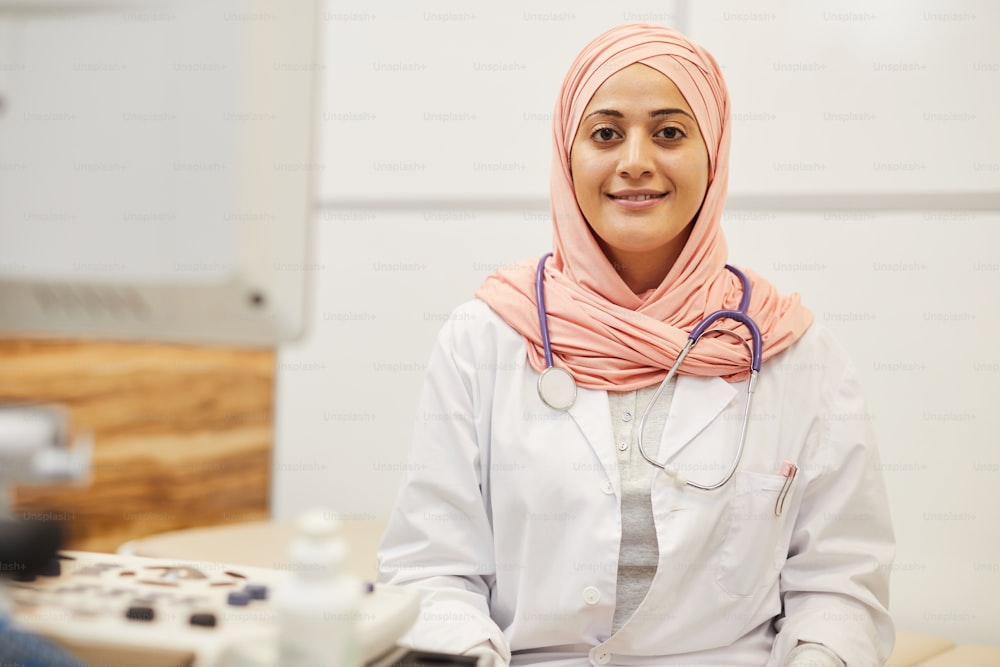 Waist up portrait of Middle-eastern woman wearing headscarf smiling at camera while posing in doctors office, copy space