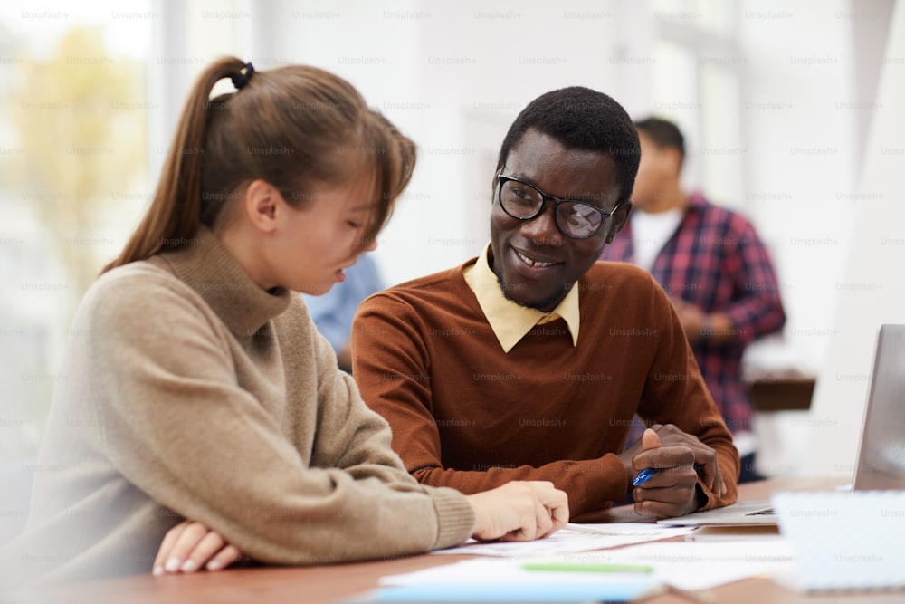 Portrait of smiling African-American student sitting at desk in college and enjoying class with female classmate, copy space