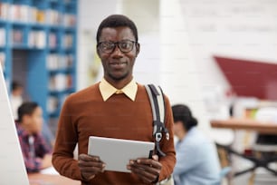 Waist up portrait of smiling African-American student looking at camera while standing in library of modern college, copy space