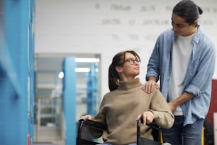Portrait of young couple, woman in wheelchair, looking at each other and smiling while standing by shelves in library