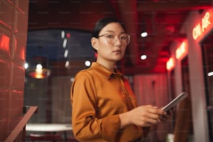Cintura para cima retrato da empresária asiática moderna segurando tablet e olhando para a câmera enquanto posa ao ar livre, espaço de cópia