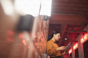 Retrato da vista lateral da jovem empresária asiática segurando o tablet enquanto se apoia na parede de tijolos ao ar livre, espaço de cópia