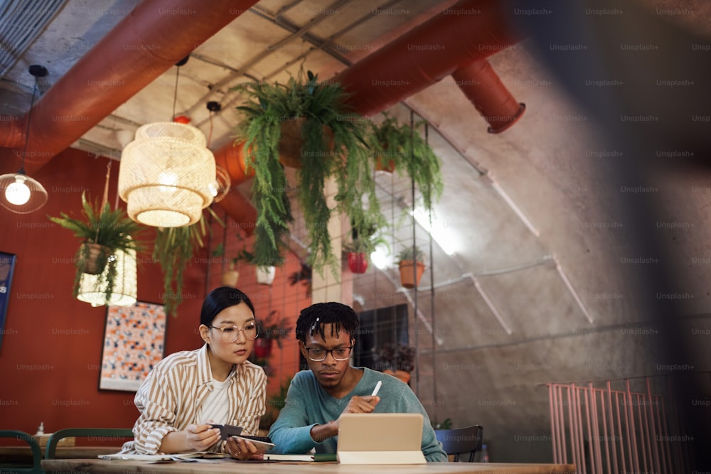 Retrato grande angular de dois estudantes contemporâneos trabalhando juntos no projeto enquanto estão sentados à mesa no café, espaço de cópia