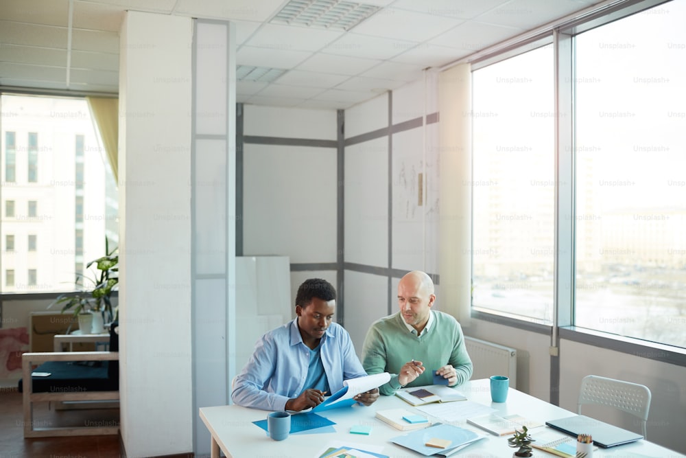 Wide angle view of African-American young man consulting with mature manager while sitting at table in office, copy space